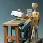 Carved painted wooden automaton. A bald man leans back in a chair. In one hand he holds a comic. The other hand is lifting a white cover revealing a green banana on a table.