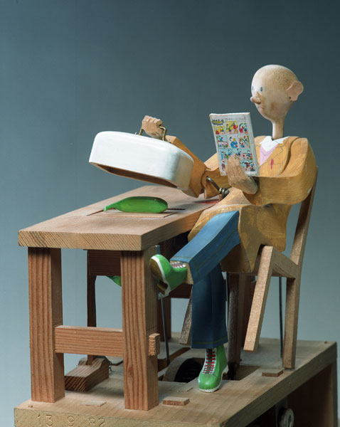 Carved painted wooden automaton. A bald man leans back in a chair. In one hand he holds a comic. The other hand is lifting a white cover revealing a green banana on a table.