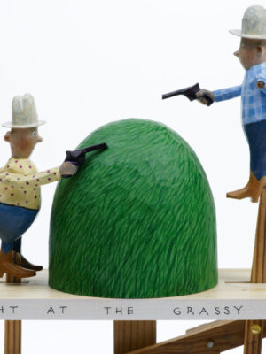 Carved wooden automata. 2 men in traditional cowboy outfits aim guns at each other either side of a grassy knoll. They alternate moving up and down.