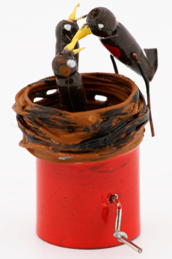 A miniature birds nest with 2 baby robins inside and an adult robin feeding them. Mounted on a red base with wire handle