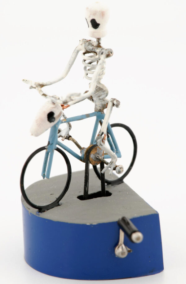A tiny skeleton rides a blue bicycle. The handlebars are made with a horned animal skull. It is mounted on a blue and grey base with metal handle.