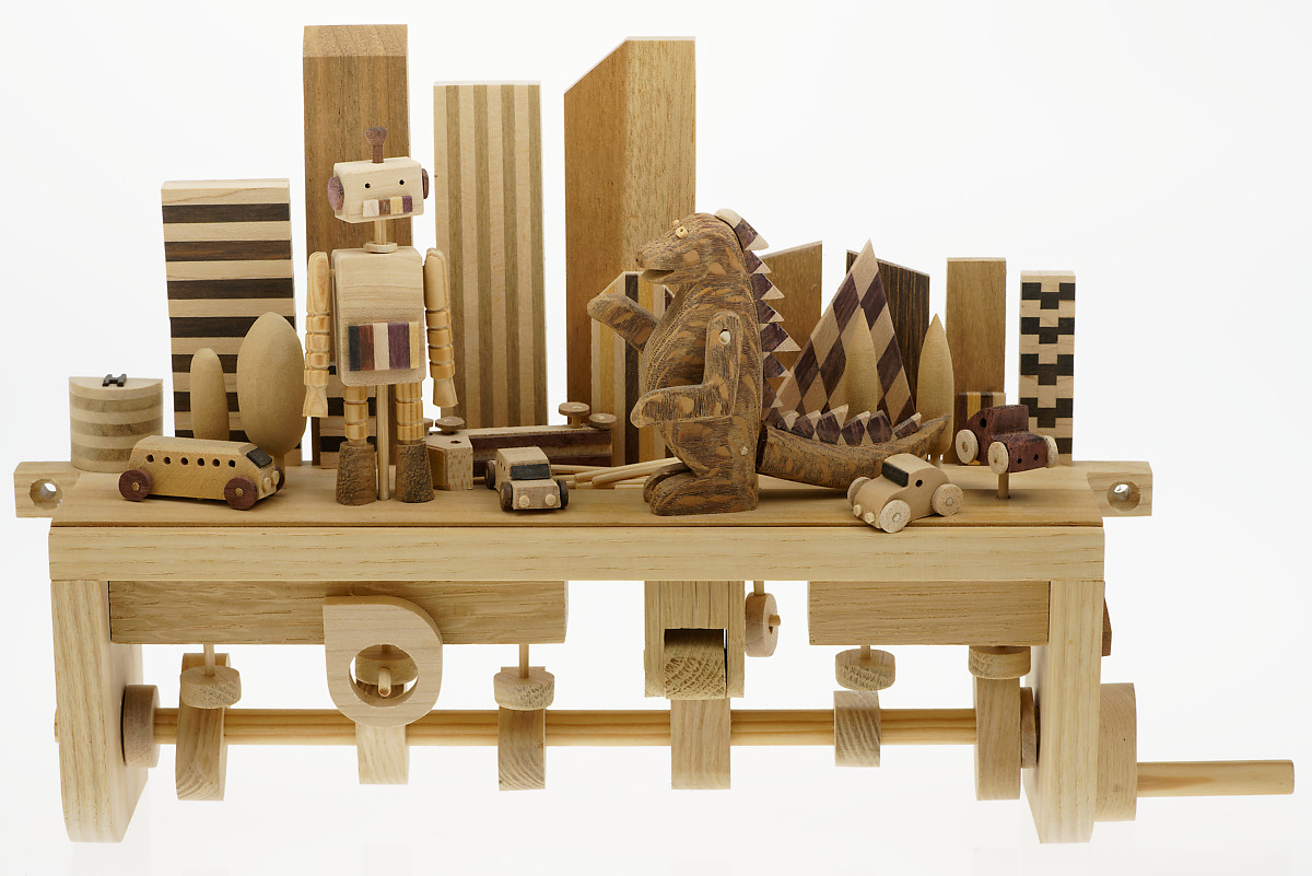 Handmade wooden automaton featuring a city skyline with a large robot and godzilla in front, there are also small cars and trees completing the scene
