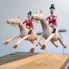 2 carved wooden horses mid leap, they are being ridden by men in red jackets and black top hats