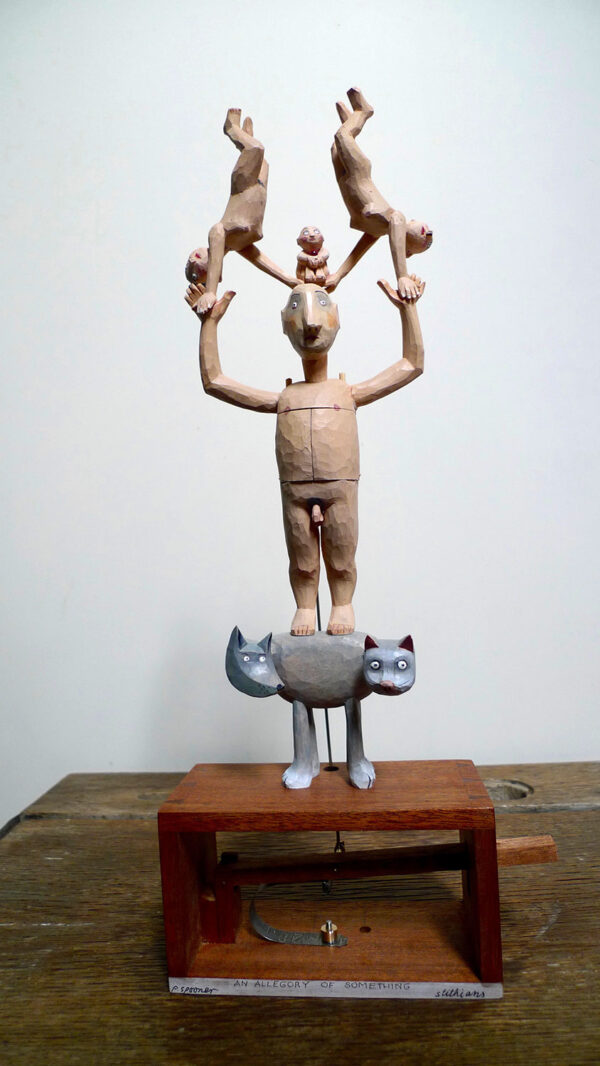 Carved painted wooden automata. A pale naked man stands on a 2 headed grey animal, one head is a cat, the other is a dog. The man holds 2 figures above his head, they are doing hand stands. A small baby sits on his head