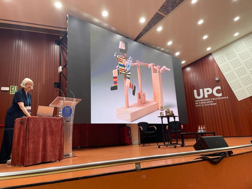 Sarah stands at a podium on the side of the stage. Peter Markeys Runner automaton is projected on the screen. It is a simple wooden man wearing brightly coloured stripy clothes in a running position.