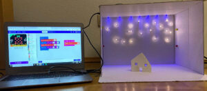 A laptop with stepped programming is attached to a white box lit with lids and a small house in the centre. It looks like a snowy scene.