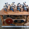 12 painted carved wooden figures sit at benches beating squares of gold. The base is filled with cogs and levers.