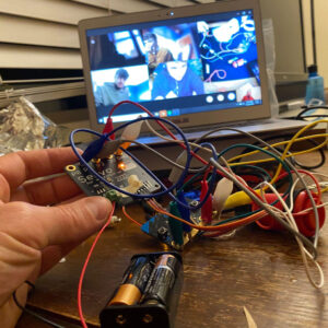 A hand holds some electronic components. There is a laptop with a multi person zoom call in the background.