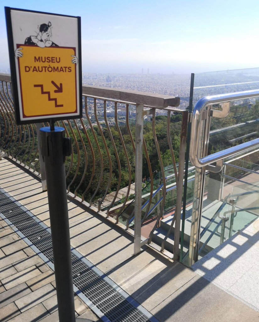 A sign reads "Museu d'AutÃ²mates". It points down some steps. A panoramic view of Barcelona in the haze is in the background.