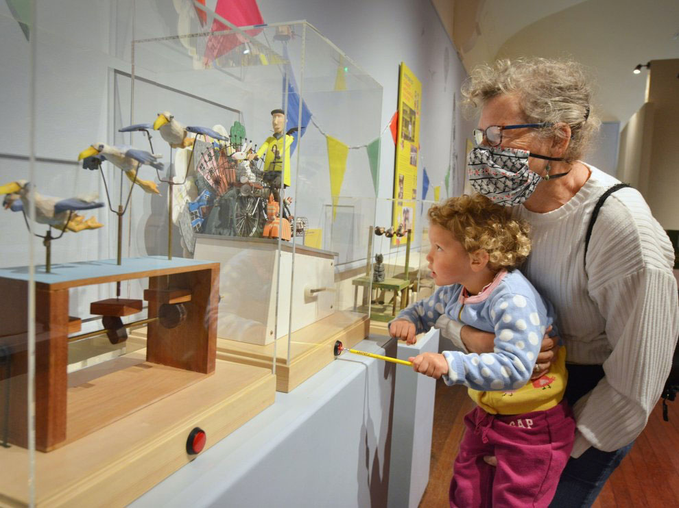 A woman holds up a small child to push a button to operate an automata of a man on a bike wearing yellow jacket. In the foreground another machine features flying birds.