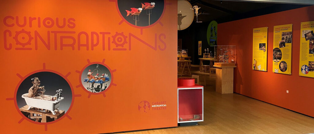 An exhibition space. A bright orange wall with text that reads 'curious contraptions' There are 3 circles containing pictures of automata. To the right you can see plinths with exhibits, it is very inviting.
