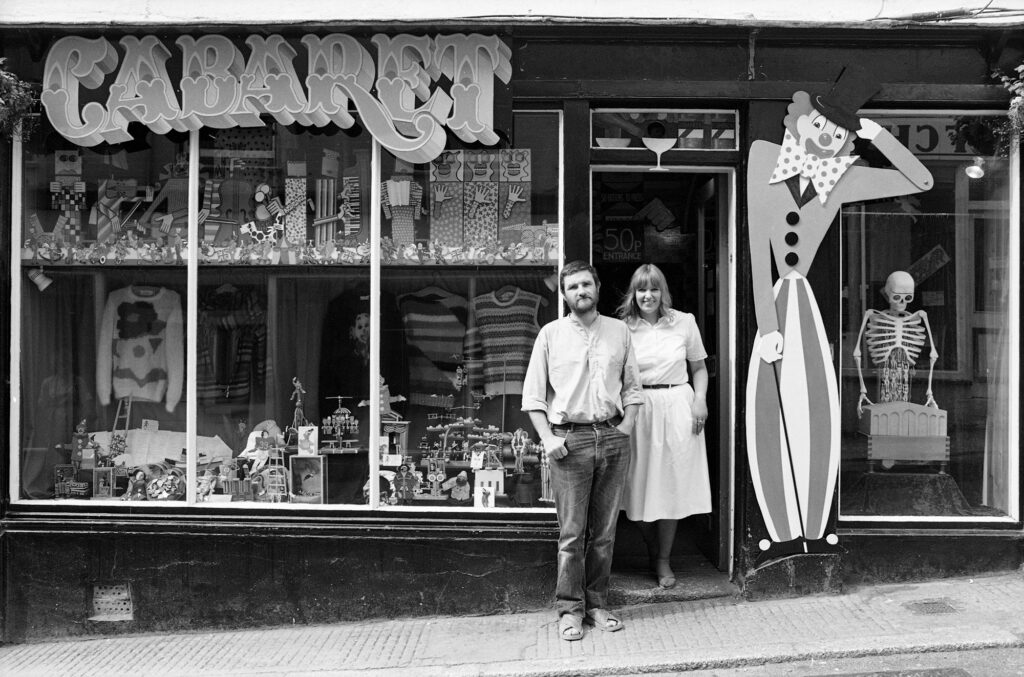 black and white image of 2 people standing in a shop door, the sign says CABARET, there is a painted clown on the right and the shop window on the left is filled with small artworks.