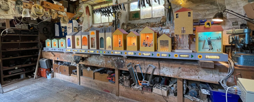 A workbench filled with small wooden boxes with roof shaped tops. Each has a large window with a mechanical scene inside. The workshop walls are filled with tools and materials.