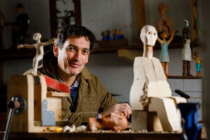 Carlos Zapata smiles at the camera. he is in a busy workspace surrounded by his carved and painted figures.