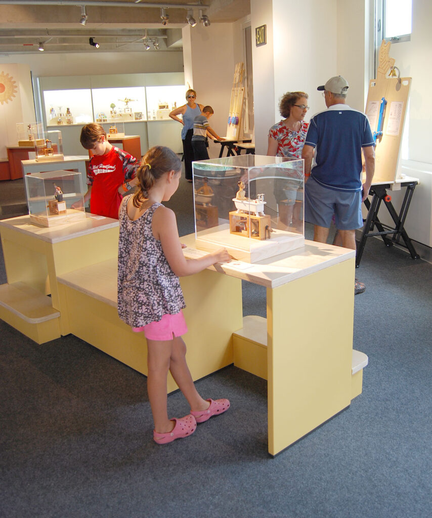 A girl press a button to activate an automaton of a man in a bath eating spaghetti. Behind her more visitors are interacting with exhibits.