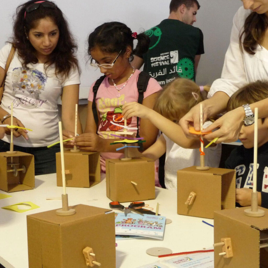 A table filled with cardboard boxes containing cam mechanisms. Women and children are constructing and playing with the pieces.