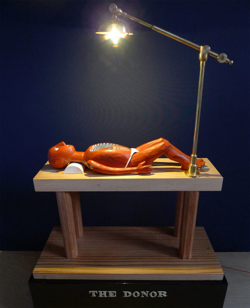 A carved wooden figure lying on the table with their legs bent. They are painted red and their organs are exposed. A lamp looms over the table.