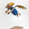 A carved painted wooden flying angel in a blue dress with golden hair and brass wings. Her arms are stretched forward. She is suspended by a bendy wire above a block of wood.