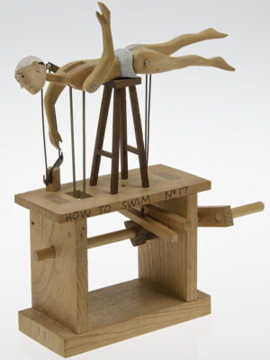 A carved wooden figure in swimming cap and stripy pants is balance horizontally on a stool. When the hanlde is turned his legs kick and arms rotate as if he is swimming.