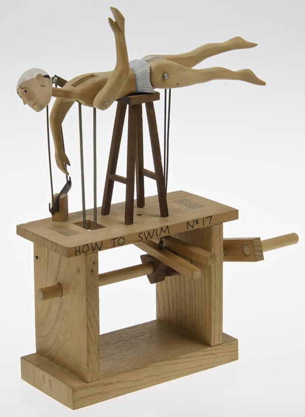A carved wooden figure in swimming cap and stripy pants is balance horizontally on a stool. When the hanlde is turned his legs kick and arms rotate as if he is swimming.