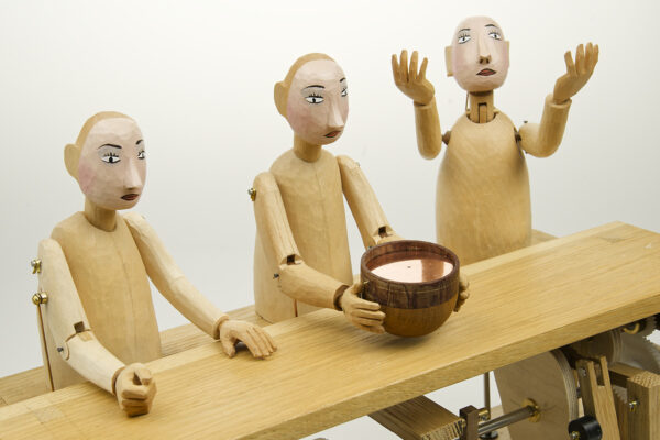 3 carved figures with painted faces sit at a table demonstrating the properties of solid, liquid and gas.
