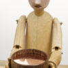 A carved wooden figure with painted face holds a bowl with a titlting copper disc inside.